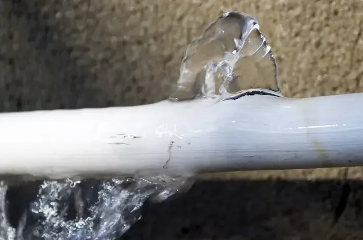 Water sprays through a split in a white plastic supply pipe